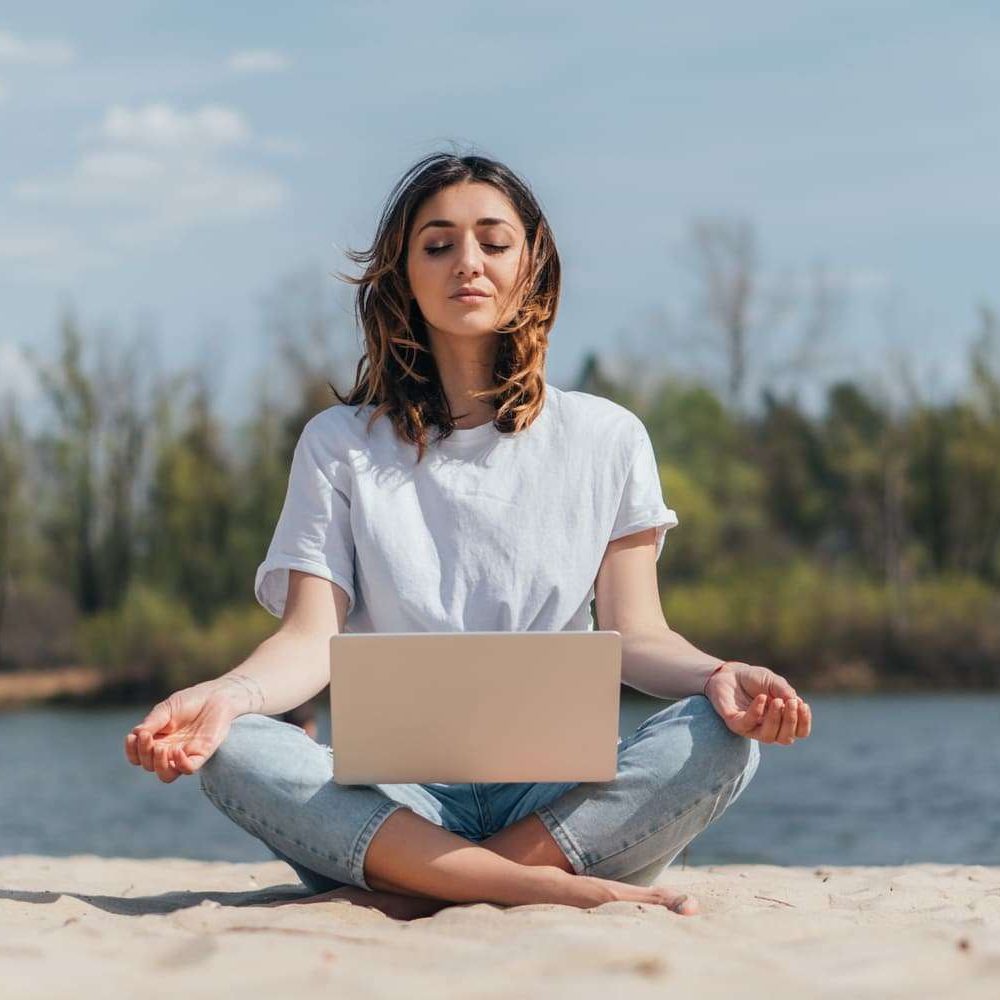 attractive freelancer with closed eyes meditating on sand near laptop