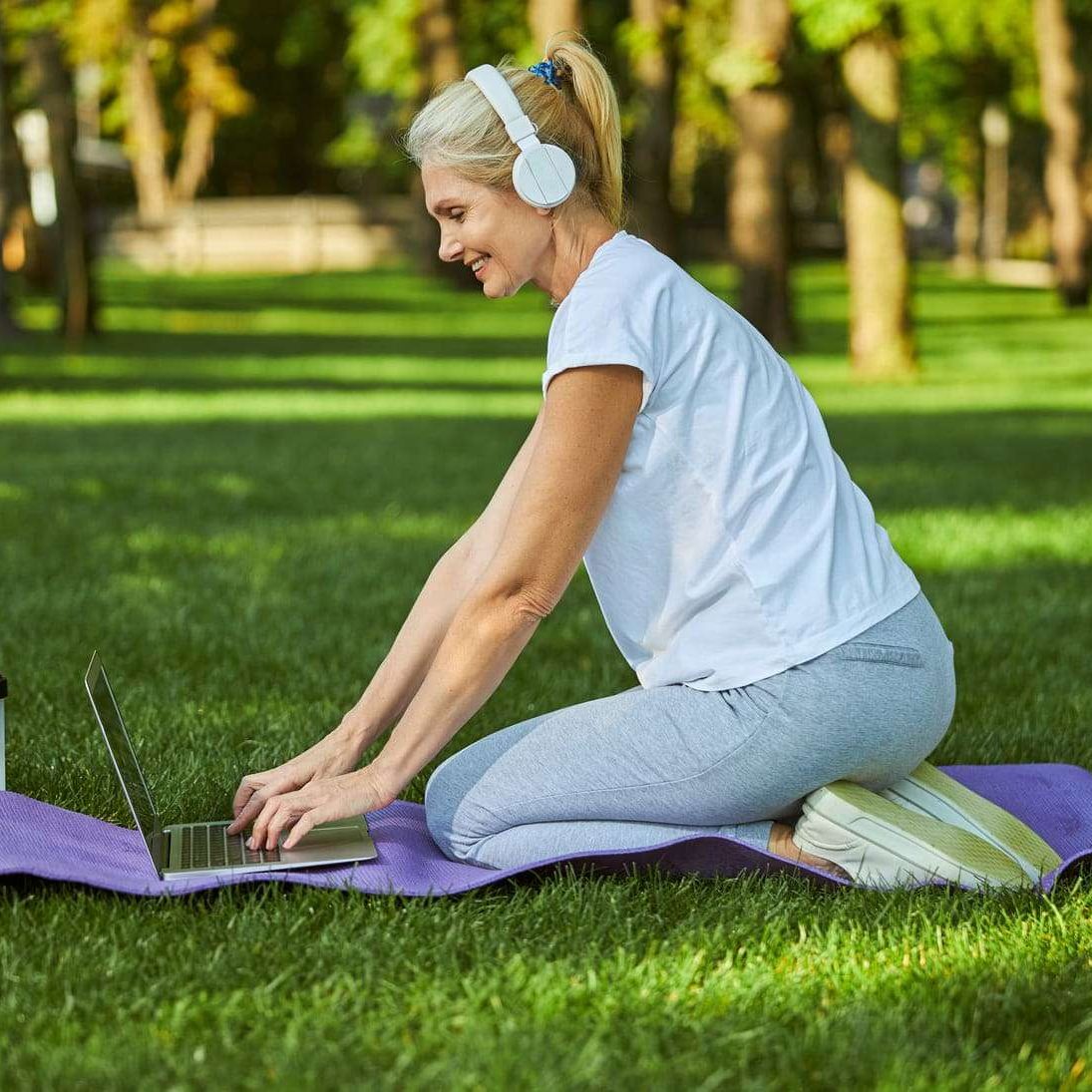 Beautiful lady in sportswear typing on laptop keyboard and smiling while sitting on yoga mat on the grass
